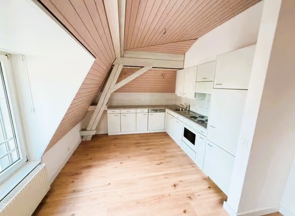 Bright, modern kitchen with white cabinets, wooden flooring, and built-in appliances including a stove and oven. The room features a slanted ceiling with exposed wooden beams and large windows allowing natural light.