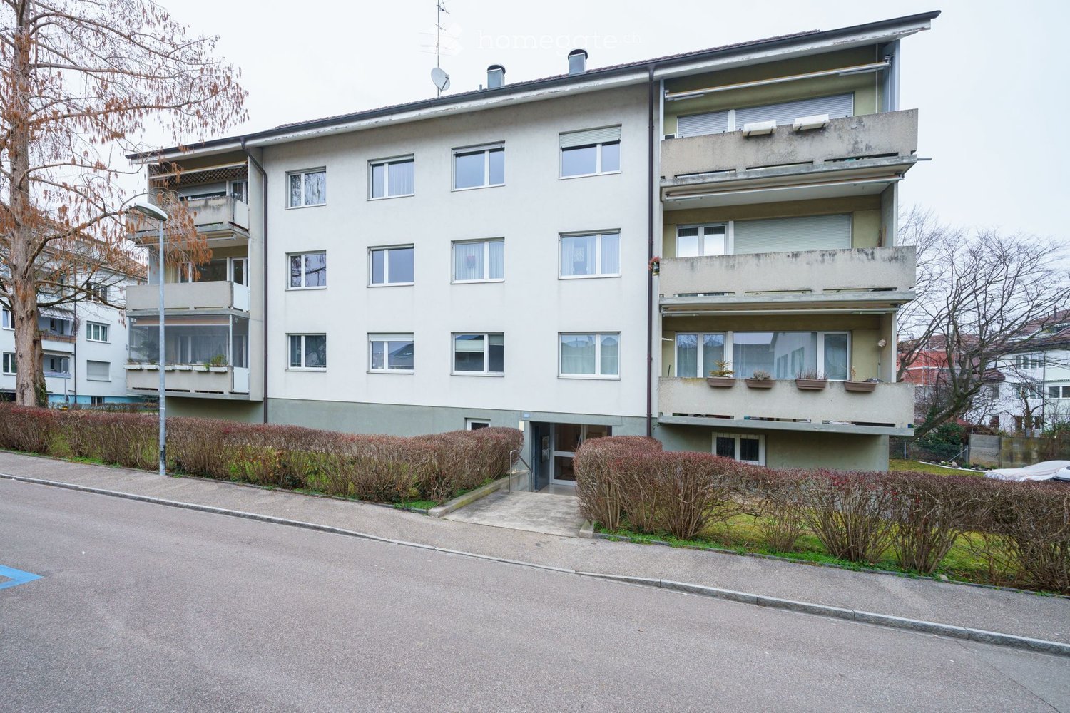 Four-story residential building with balconies, multiple windows, and a concrete facade. The building is surrounded by low hedges and a paved driveway.