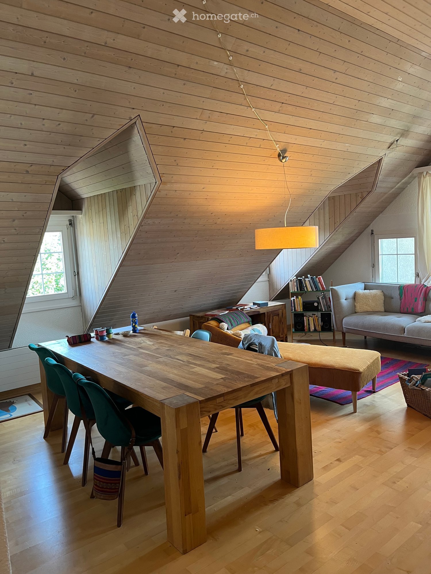 Bright, wooden-floored room with a large wooden dining table, green chairs, a sofa, a small bookshelf, and a hanging lamp.