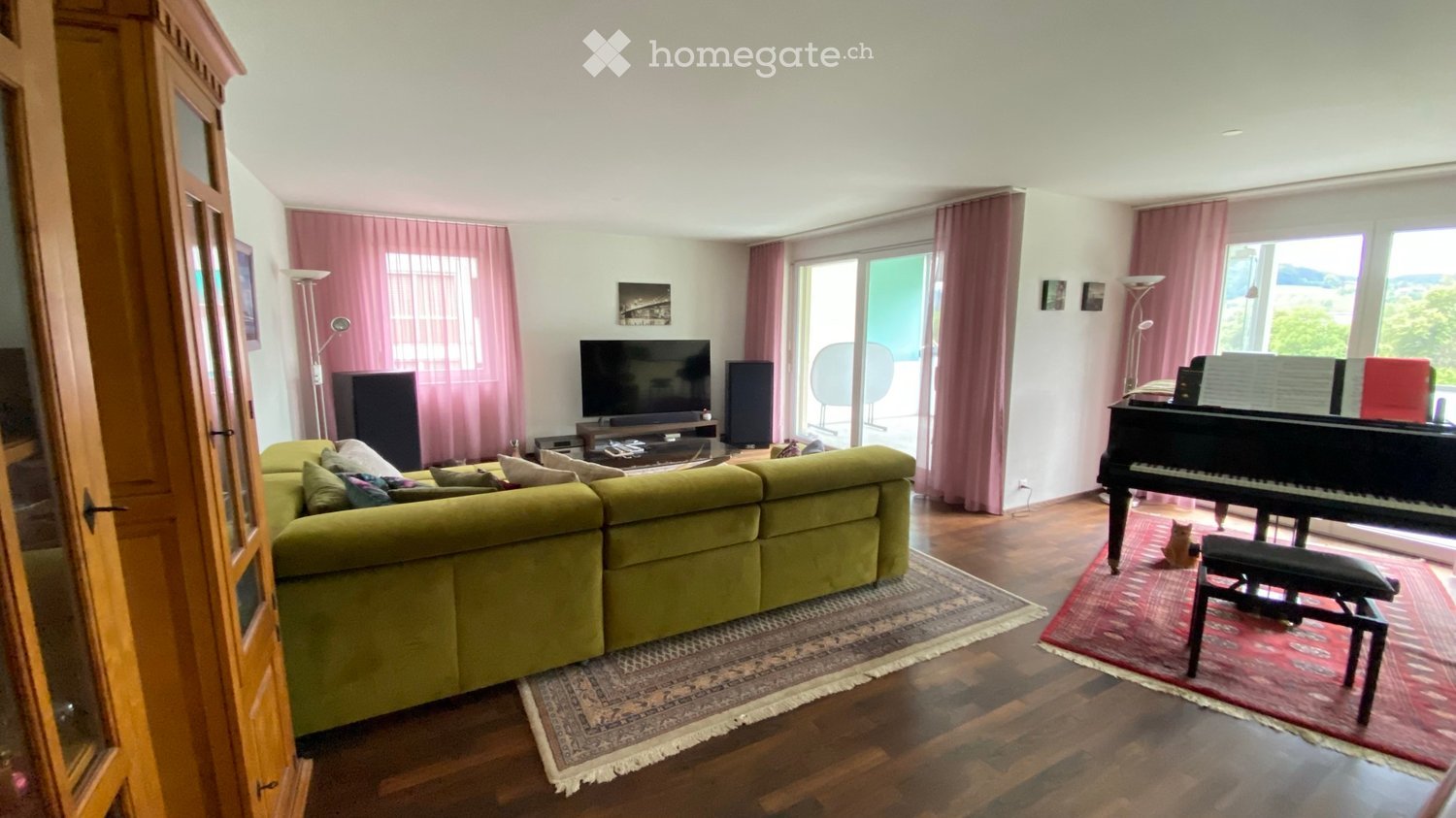 Bright living room with a green sectional sofa, a grand piano with a bench, a television on a stand, and large windows with pink curtains. The room features wooden flooring and a patterned rug.