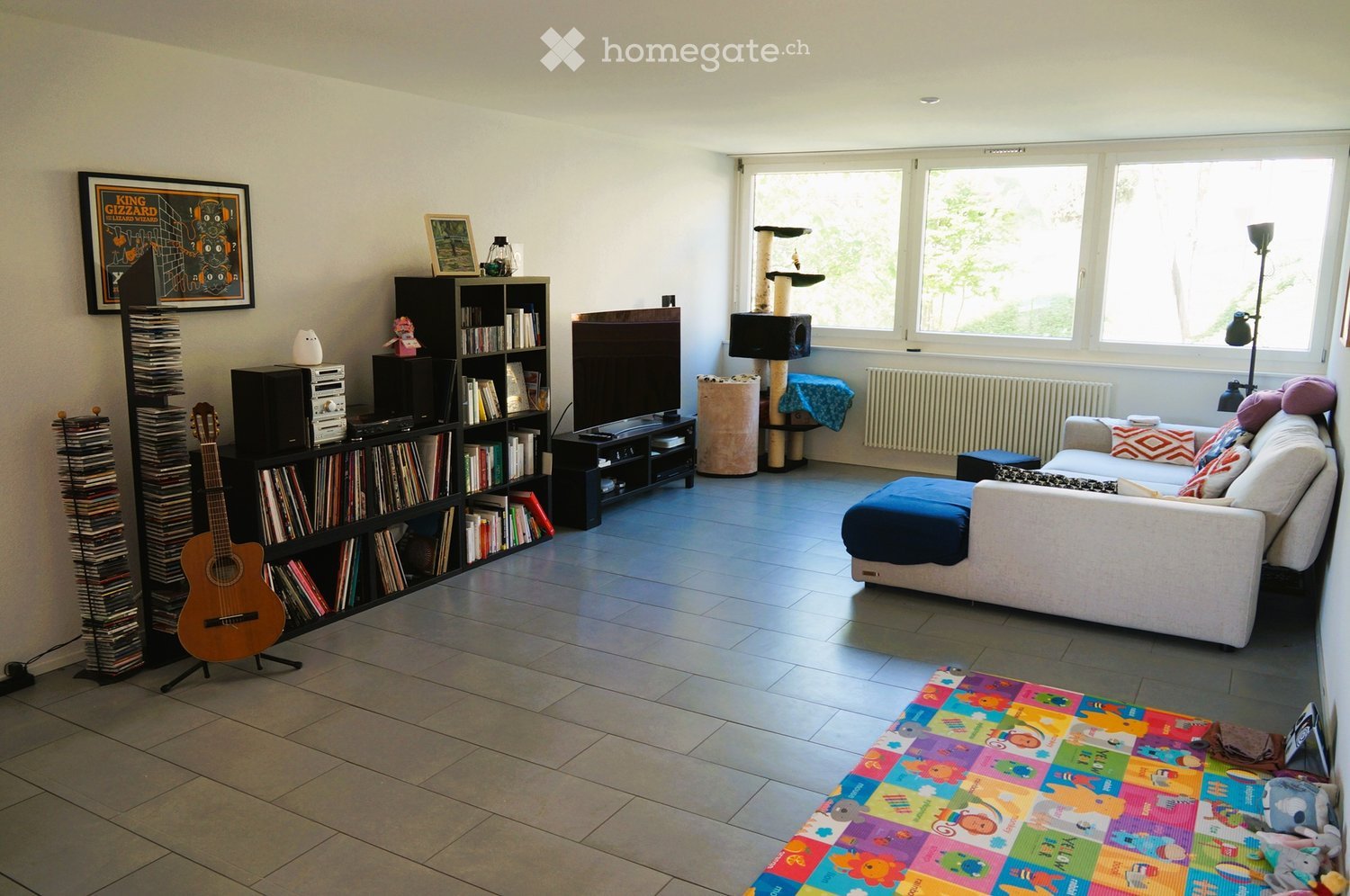 Bright living room with large windows, white walls, and tiled floor. Features a white sofa with colorful cushions, a blue ottoman, a coffee table with toys, a bookshelf with books and a guitar, a cat tree, and a floor lamp.