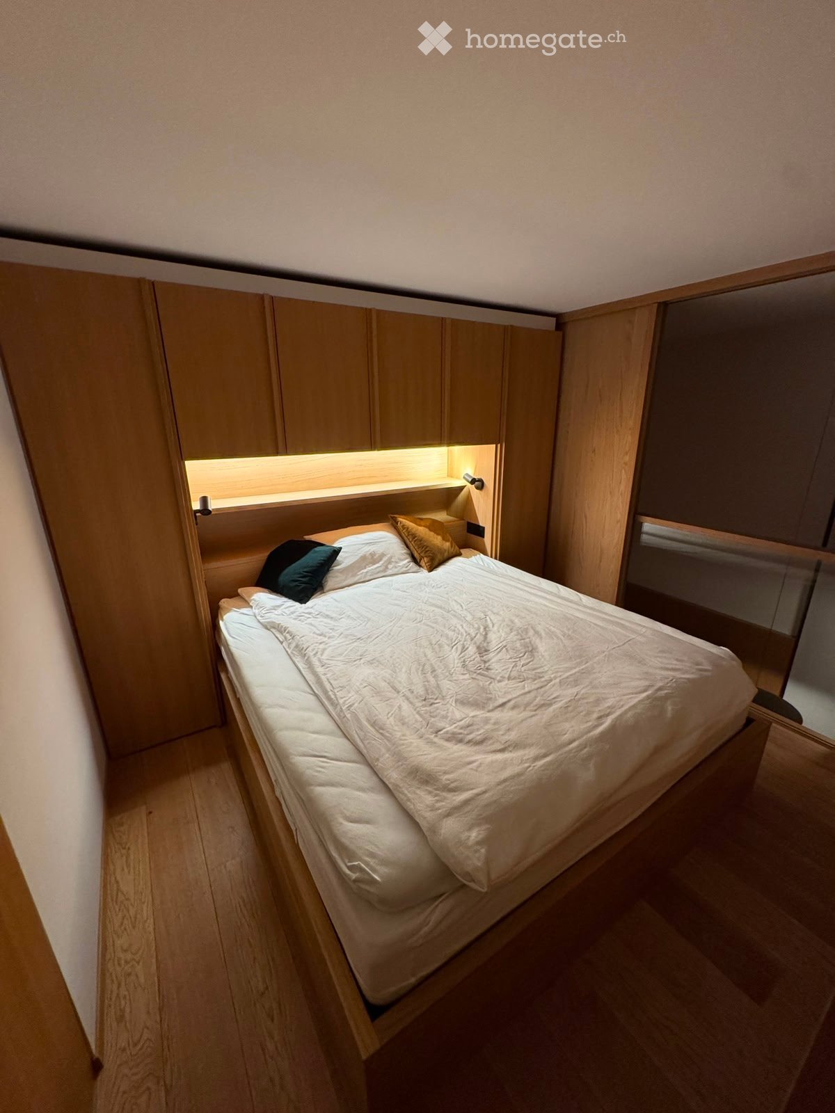 Modern, compact bedroom with a single bed, built-in wooden cabinets, and soft lighting above the bed.