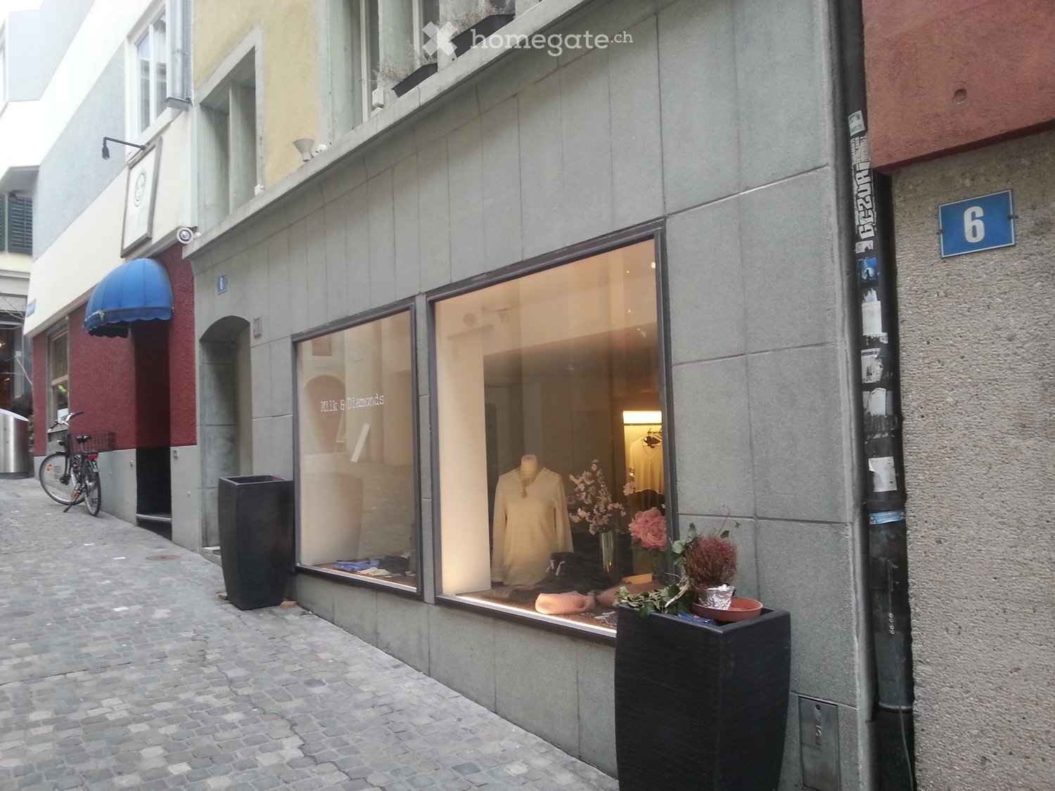 Storefront with large glass windows displaying clothing and accessories, including a mannequin, flowers, and decorative items. The building has a modern facade with grey stonework and a blue awning on the adjacent building.