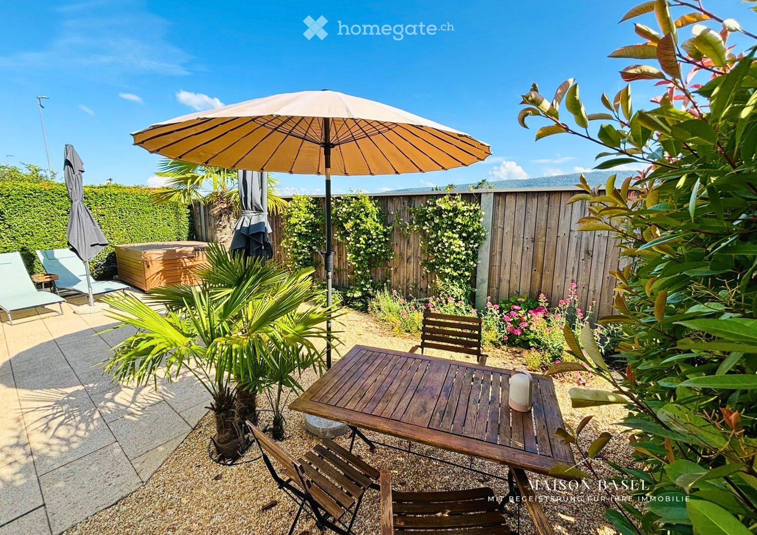 A well-maintained garden area with a wooden table, chairs, lounge chairs, a hot tub, and a large umbrella providing shade. The space is surrounded by greenery, including palm plants and flowering bushes, with a wooden fence enclosing the area.