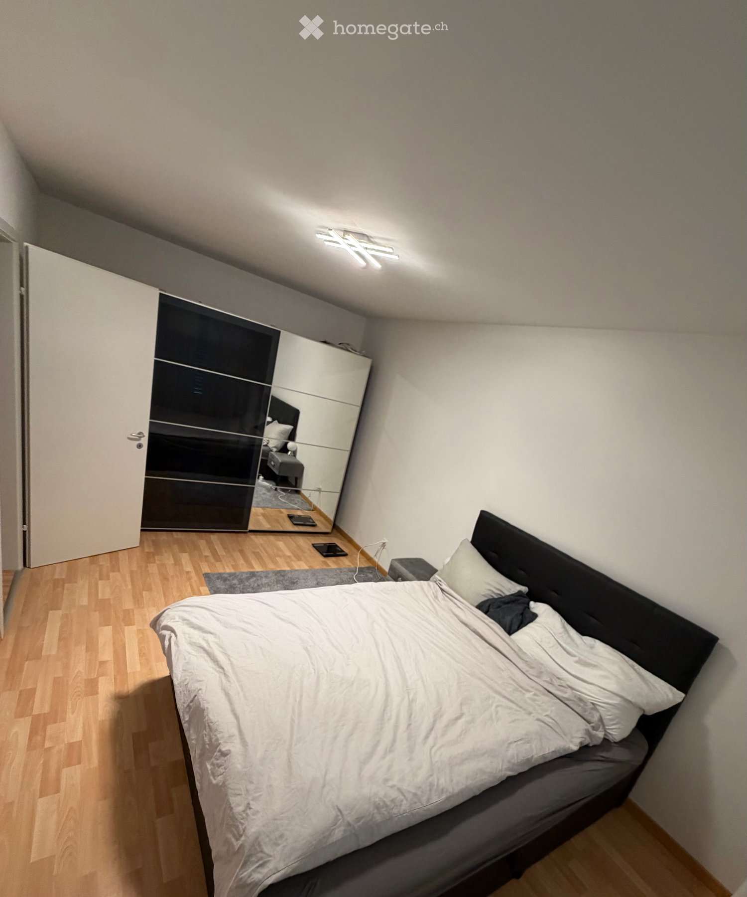 Modern bedroom with a large bed, white bedding, a black wardrobe with mirrored doors, and a small table with devices on it.