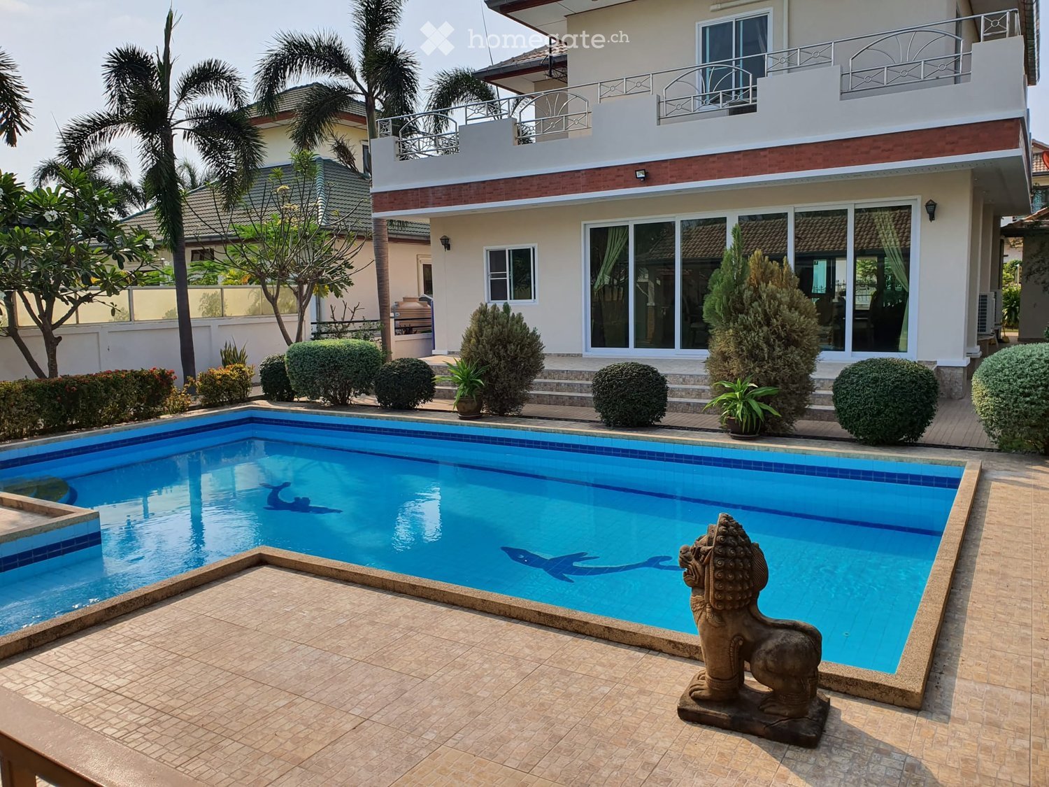 A modern two-story house with a large swimming pool in the foreground, surrounded by neatly trimmed bushes and palm trees. The pool features decorative fish silhouettes on the bottom and a stone lion statue on the deck.