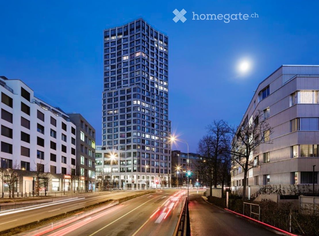 Modern high-rise building with illuminated windows, surrounded by other contemporary structures and a busy street with light trails from moving vehicles.