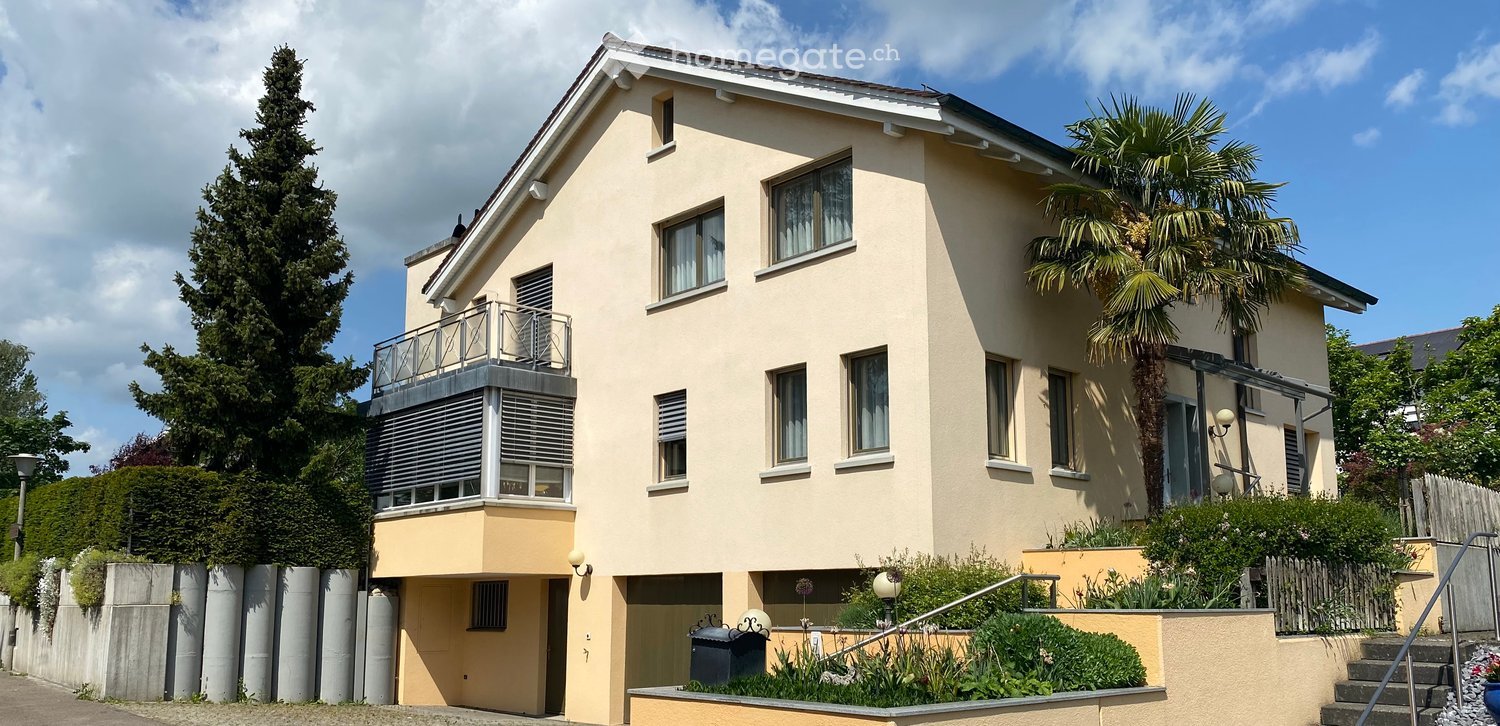 Two-story house with beige exterior, multiple windows, and a balcony on the upper floor. The house is surrounded by greenery, including a tall palm tree and shrubs.