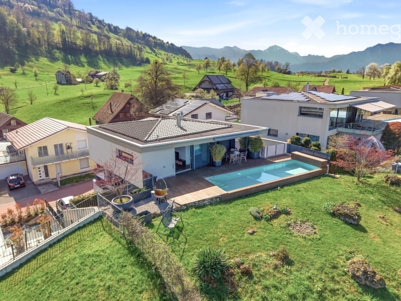 Aerial view of a modern house with a swimming pool, surrounded by a well-maintained garden and scenic mountain backdrop.