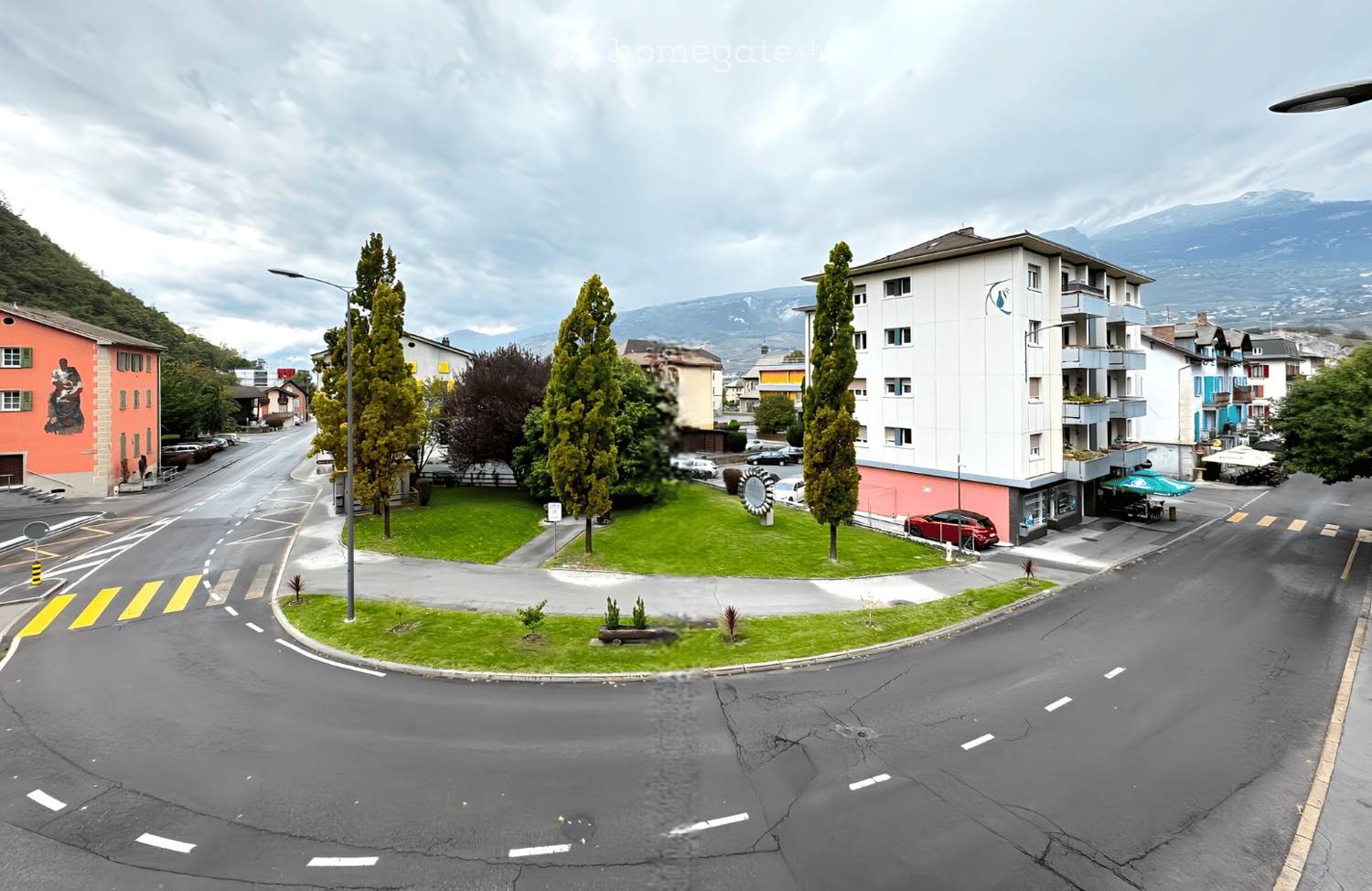 A multi-story residential building with balconies and windows, surrounded by greenery and parked cars, with a scenic mountain view in the background.