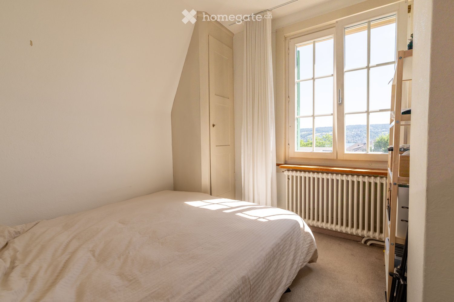 Bright bedroom with a large window offering a scenic view, featuring a neatly made bed, a radiator beneath the window, and a curtain partially drawn.