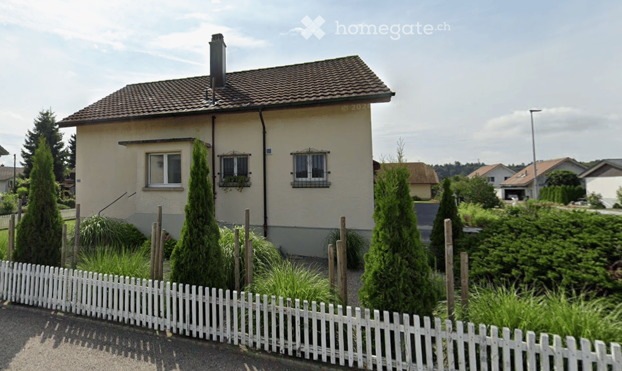 House. Single-story house with beige exterior, tiled roof, chimney, and white picket fence surrounding a garden with tall conifers and shrubs.