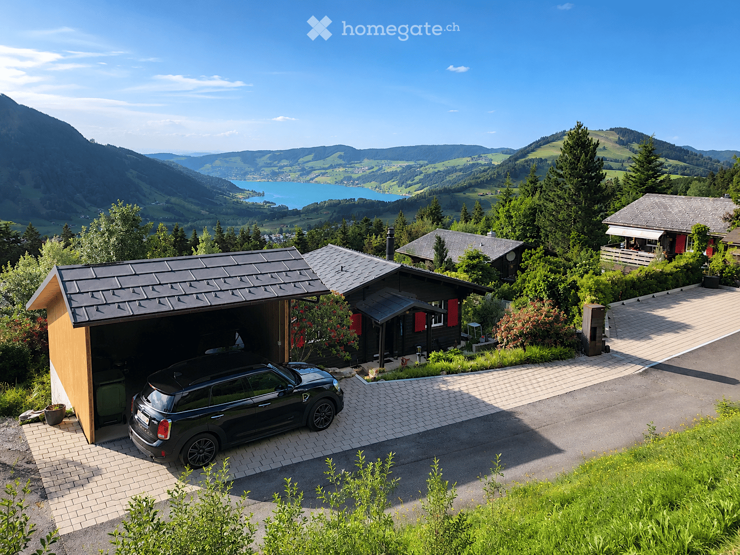 A modern two-story house with a black roof and red shutters, featuring a garage with a parked car, surrounded by lush greenery and a scenic mountain view.