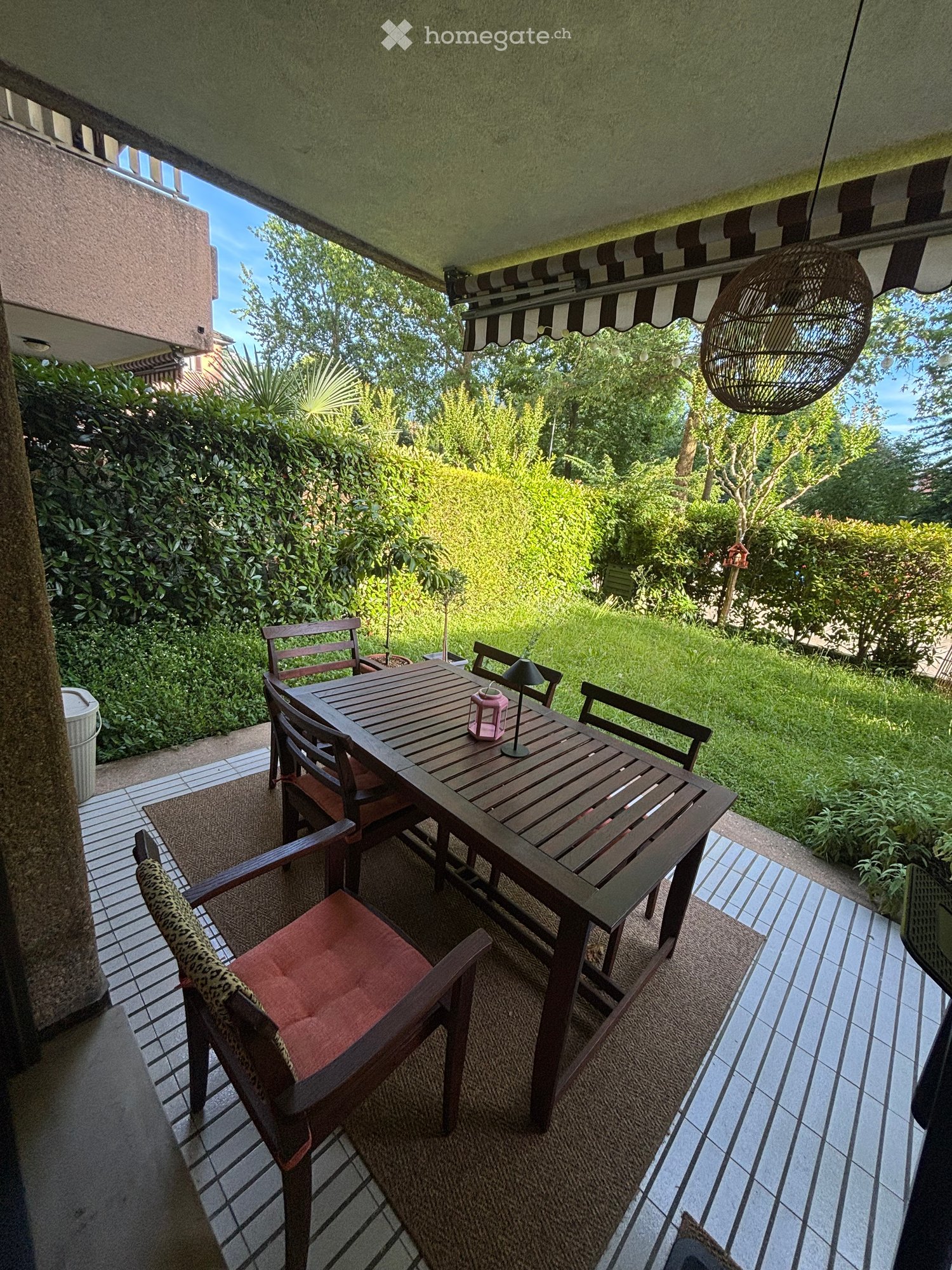 Balcony with wooden table, chairs, and a hanging lamp, overlooking a green garden.
