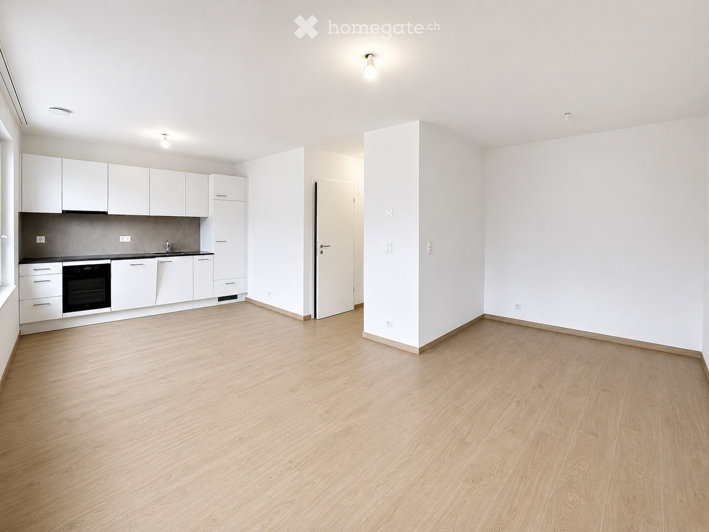 Empty room with light wooden flooring, white walls, and a small kitchen area with white cabinets and a sink.