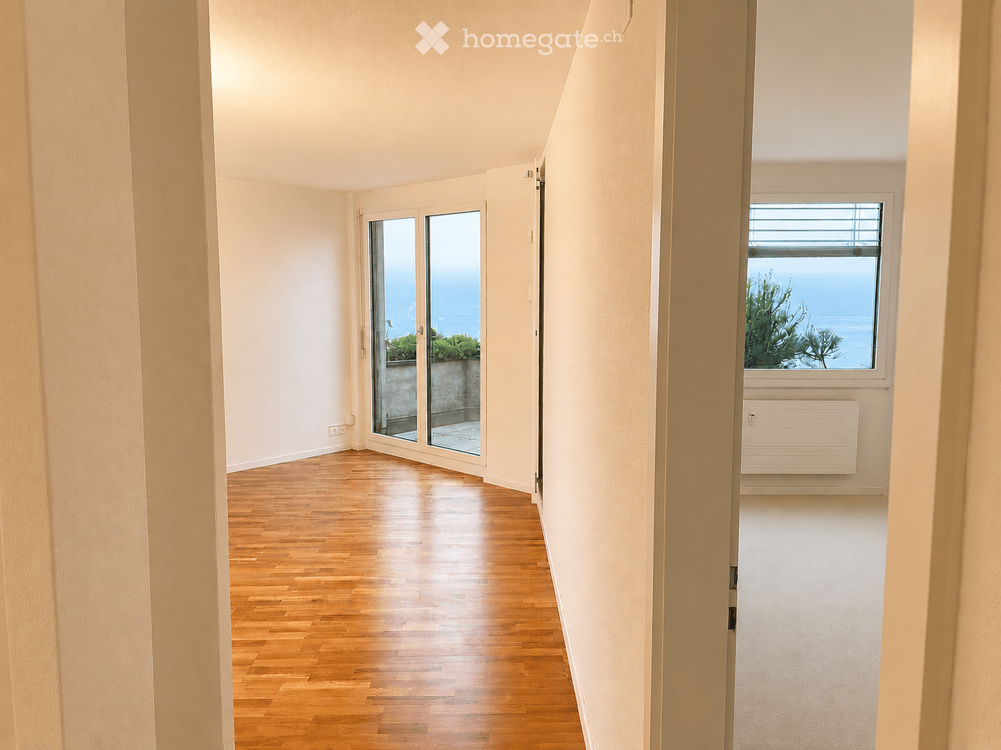 Empty hallway with wooden flooring, white walls, and large windows offering a view of greenery and water. Features a sliding glass door leading to an outdoor area.