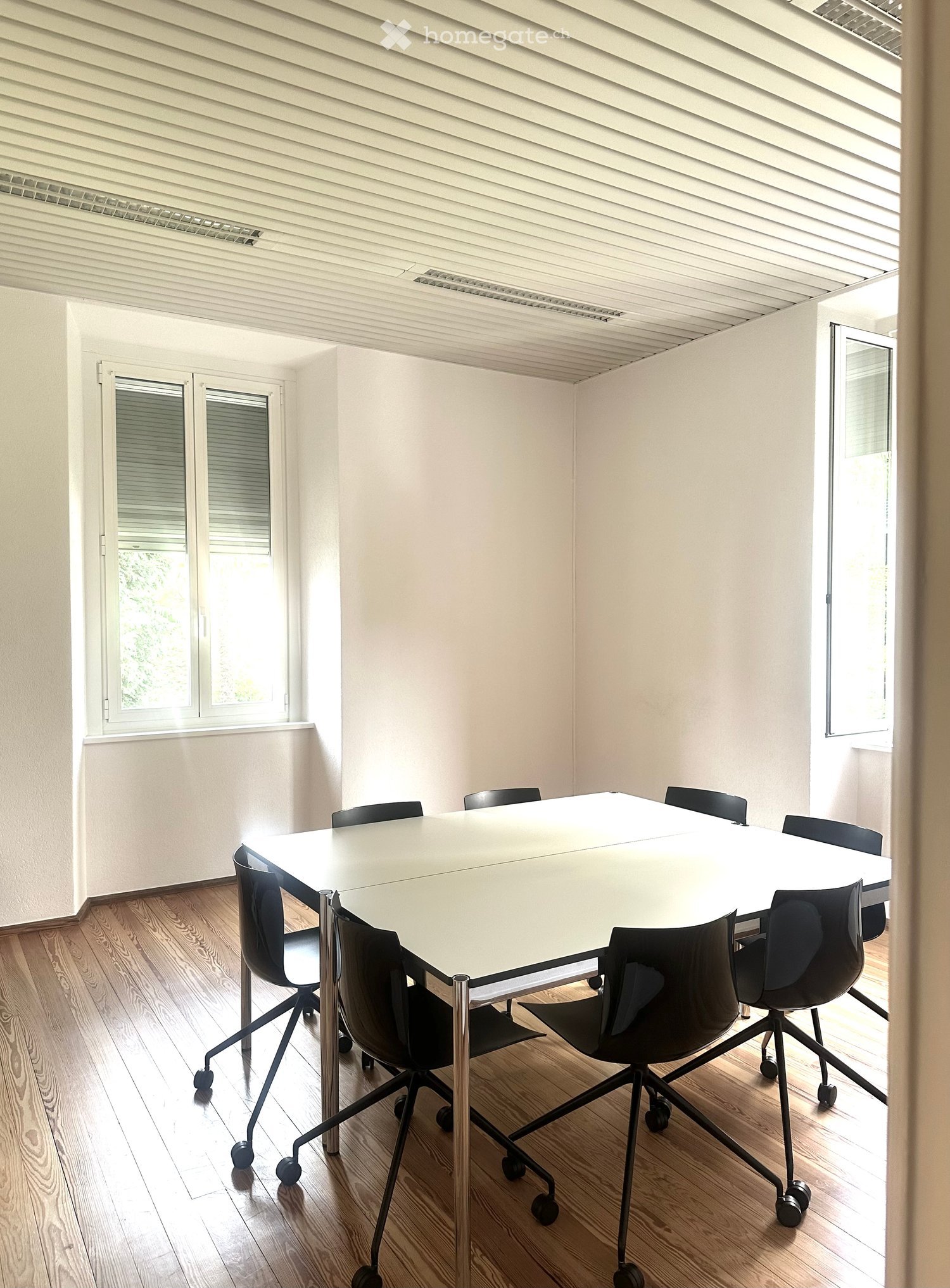 A modern, minimalist meeting room with a large white table surrounded by black office chairs, wooden flooring, and large windows with blinds.