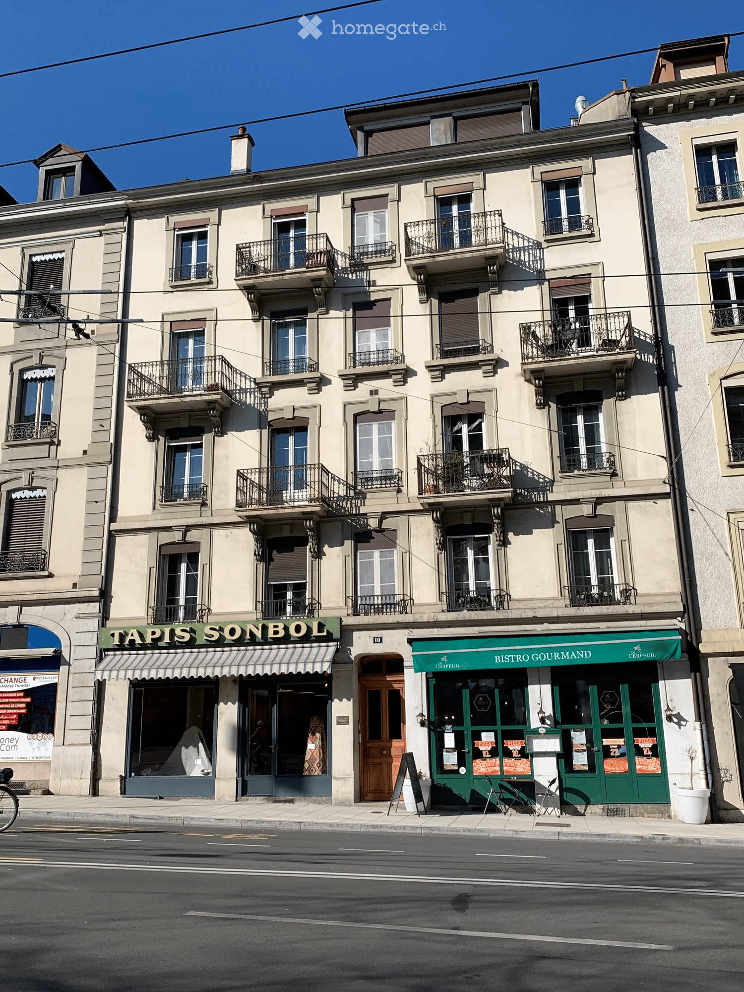 A multi-story building with beige facade, multiple windows, and balconies. Ground floor features two businesses: 'Tapis Sonbol' and 'Bistro Gourmand'.