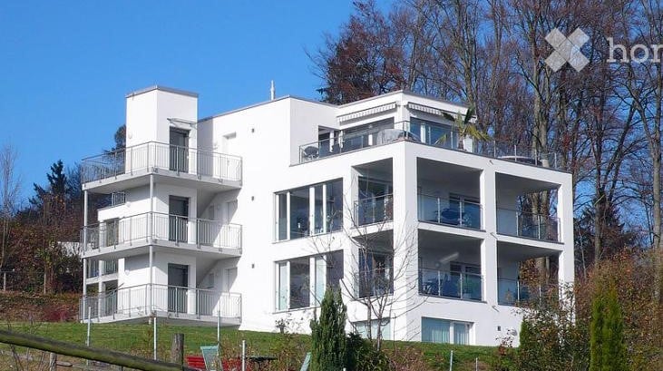 Modern white apartment building with multiple balconies and large windows, surrounded by trees.