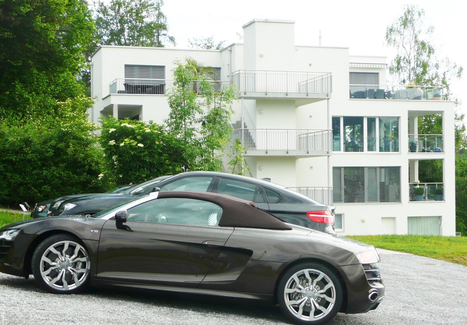 Modern white two-story building with large windows and balconies, surrounded by greenery, with several parked cars in front.