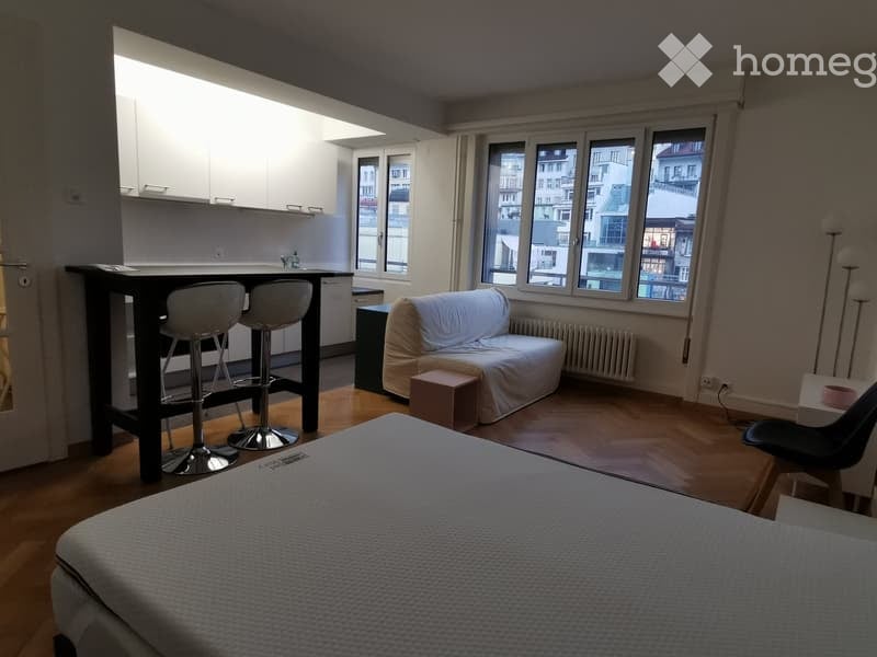 A modern, open-concept living and sleeping area with a sofa, a bed, a small table with two stools, and a view of the city through large windows.