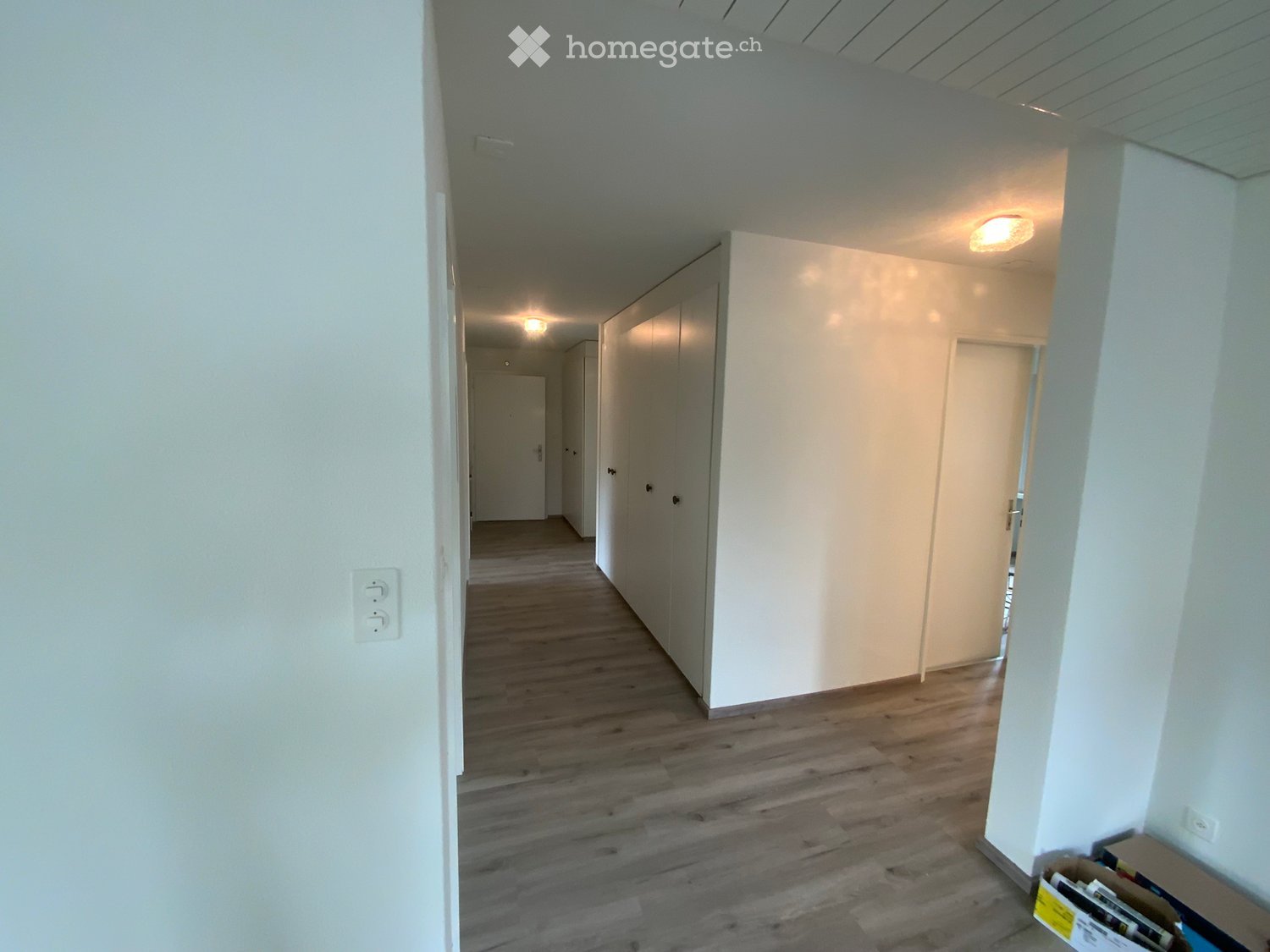 Empty hallway with wooden flooring, white walls, and several closed doors. Recessed lighting fixtures are visible on the ceiling.