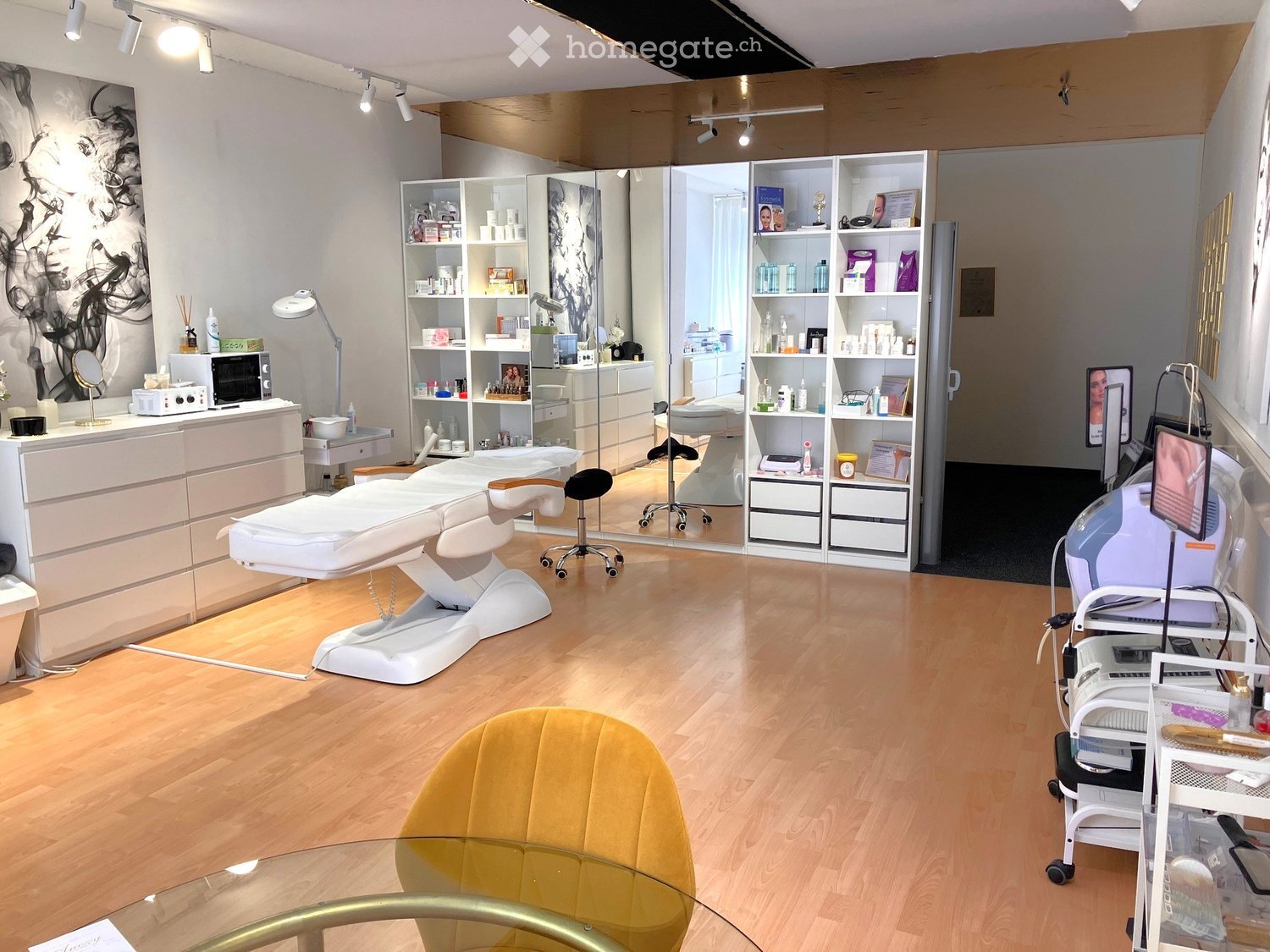 Modern, well-lit room with wooden flooring, a yellow chair, a glass table, a white treatment bed, shelves with various items, and a mirror. The space is clean and organized, with a professional aesthetic.