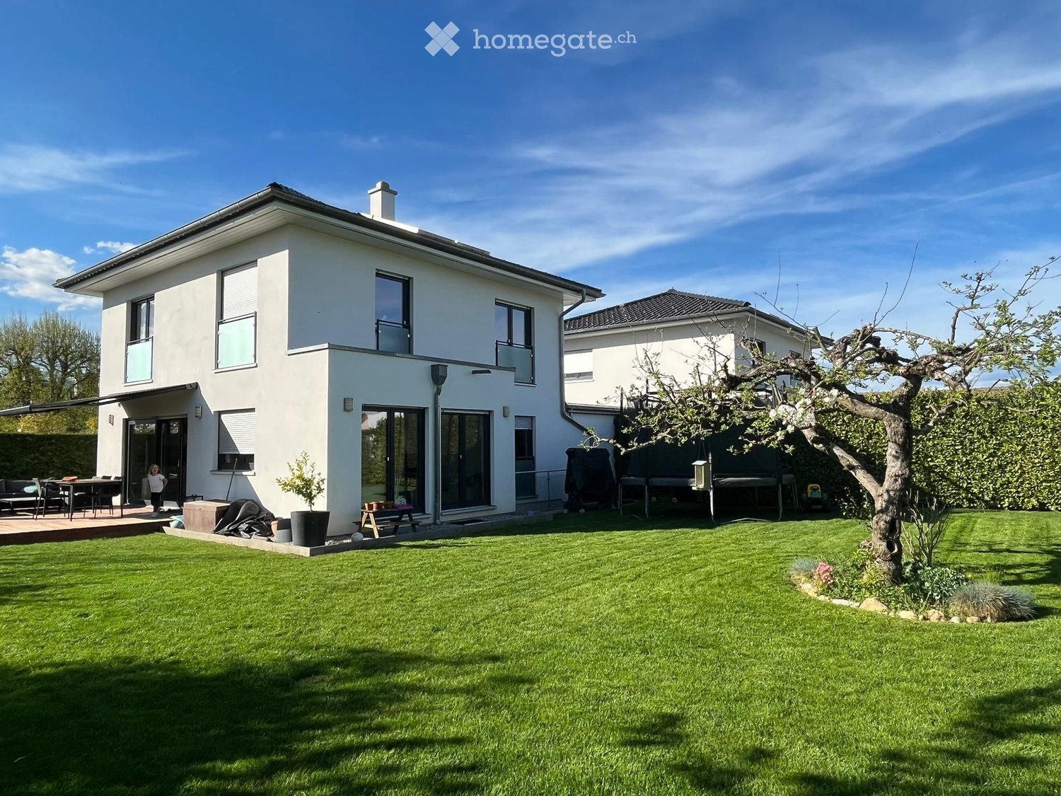 Modern two-story house with white exterior, large windows, and a spacious lawn in front. Features include a covered patio area with outdoor furniture and a small garden with trees and shrubs.