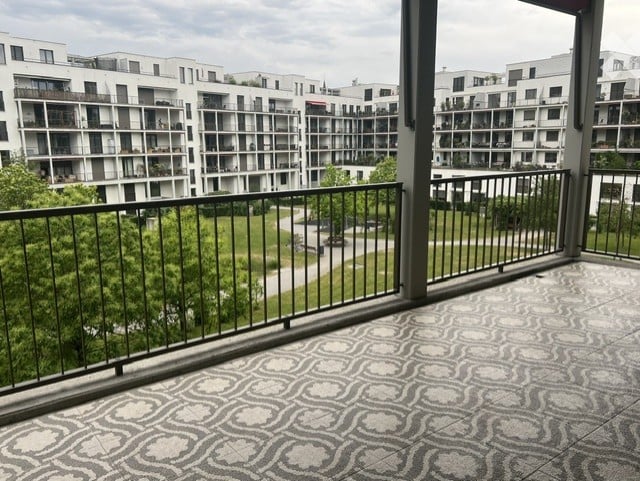 Balcony with decorative tile floor, metal railing, overlooking a green park and modern apartment buildings.