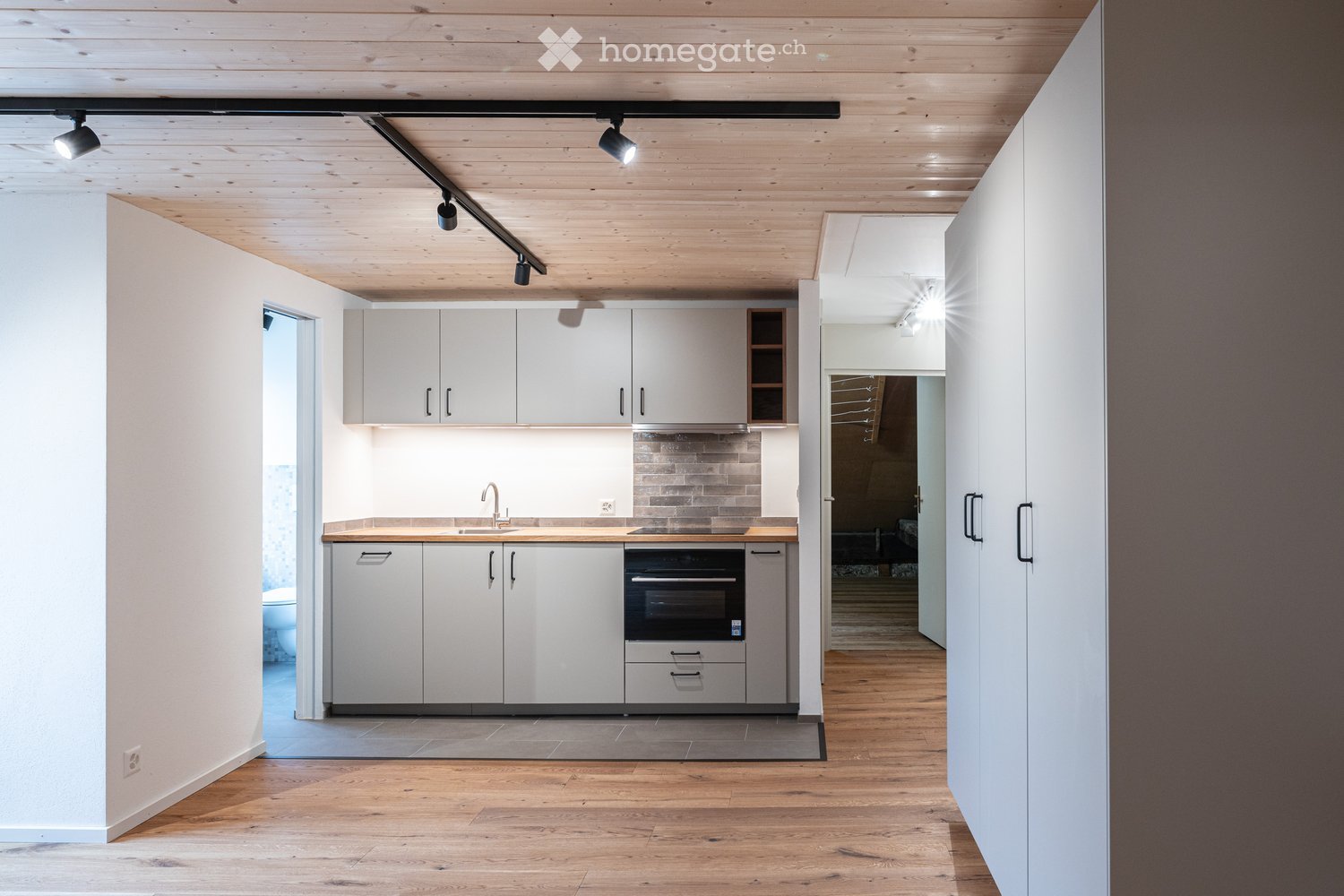 Modern kitchen with white cabinets, wooden countertops, and stainless steel appliances, including a sink and oven. The space features wooden flooring and track lighting.