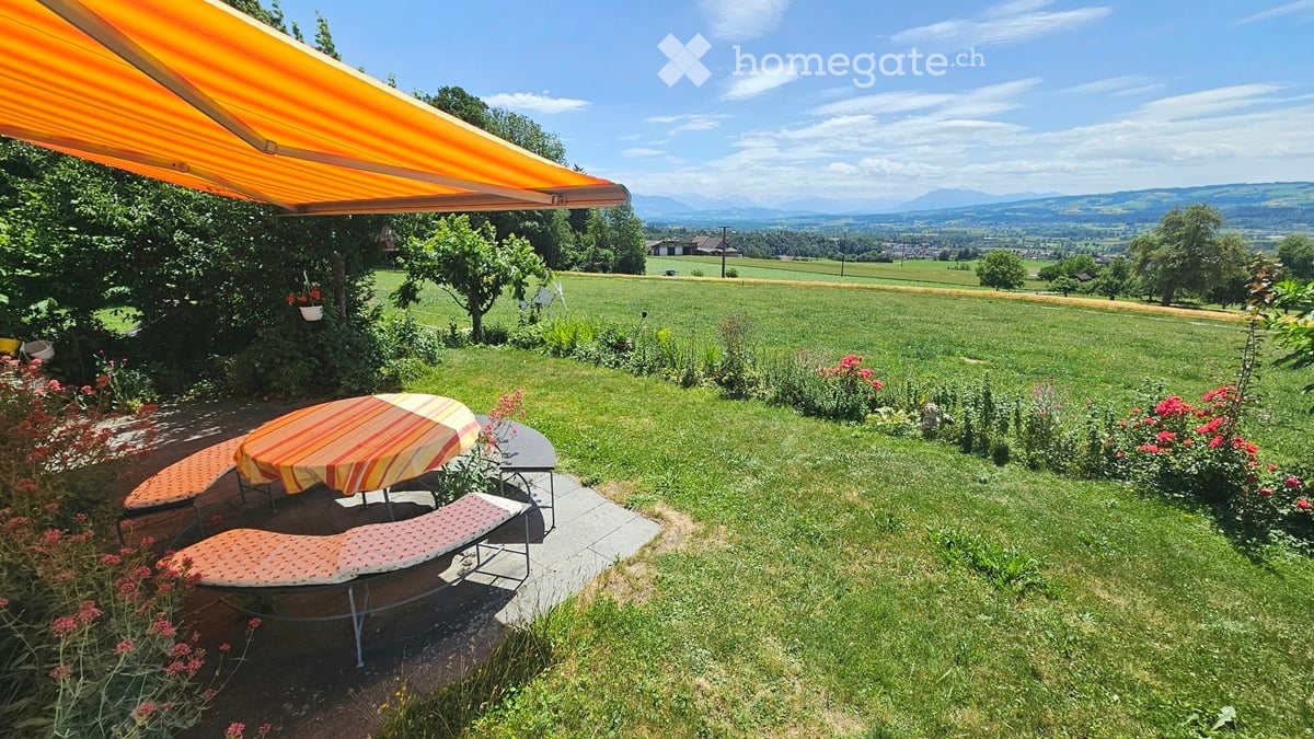 Lush garden with a patio area featuring a table, chairs, and lounge chairs under an orange awning. The background shows a scenic countryside view with fields and mountains.