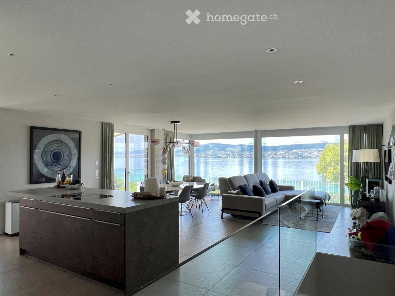 Modern open-plan living and dining area with large windows offering a scenic view, featuring a grey sofa, coffee table, dining table with chairs, and a kitchen island.