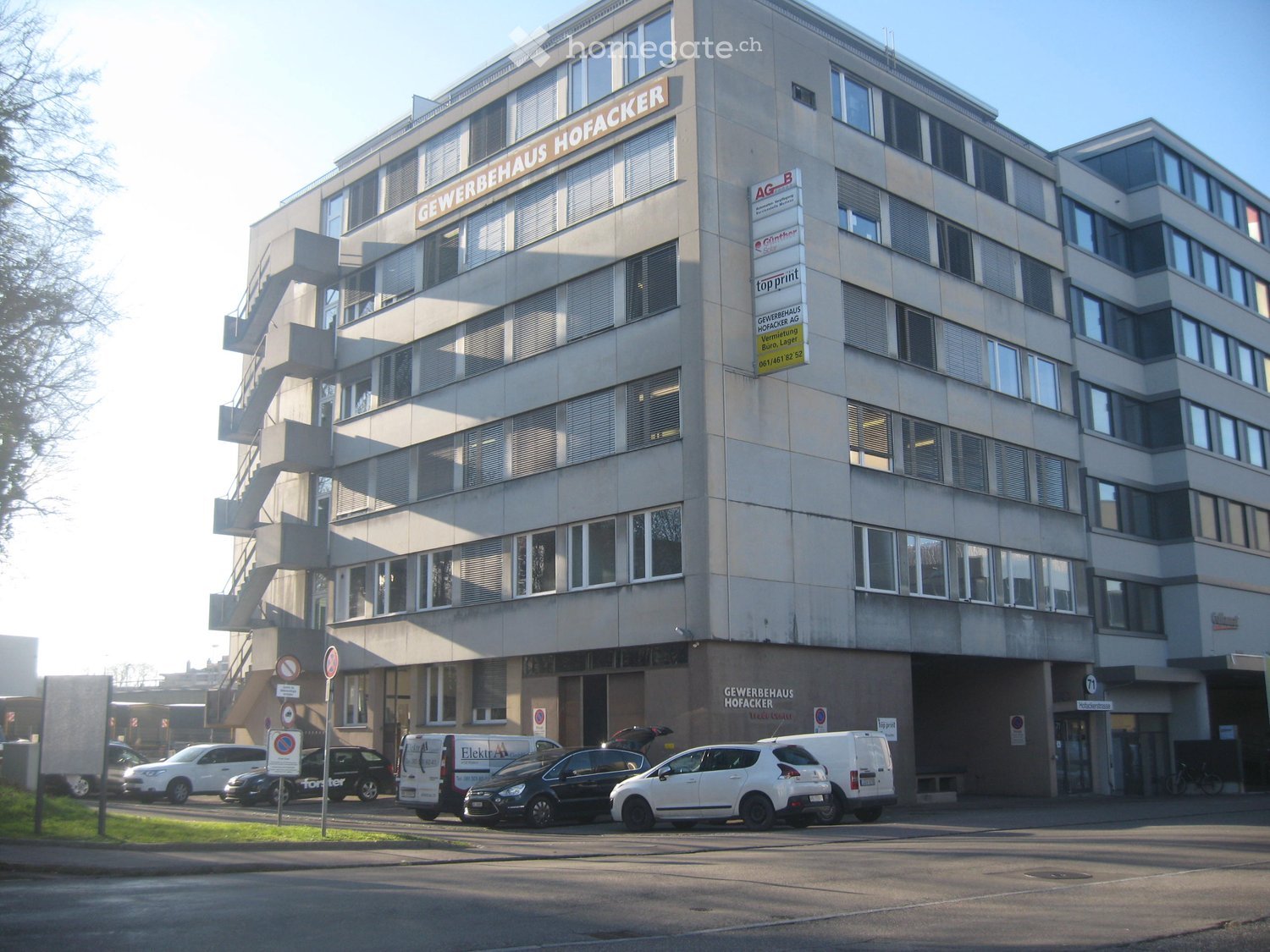 Building. Multi-story commercial building with signage for "GEWERBEHAUS HOFACKER" and several parked cars in front.