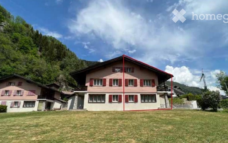 A two-story house with a red roof and white shutters, situated in a grassy area with mountains in the background.