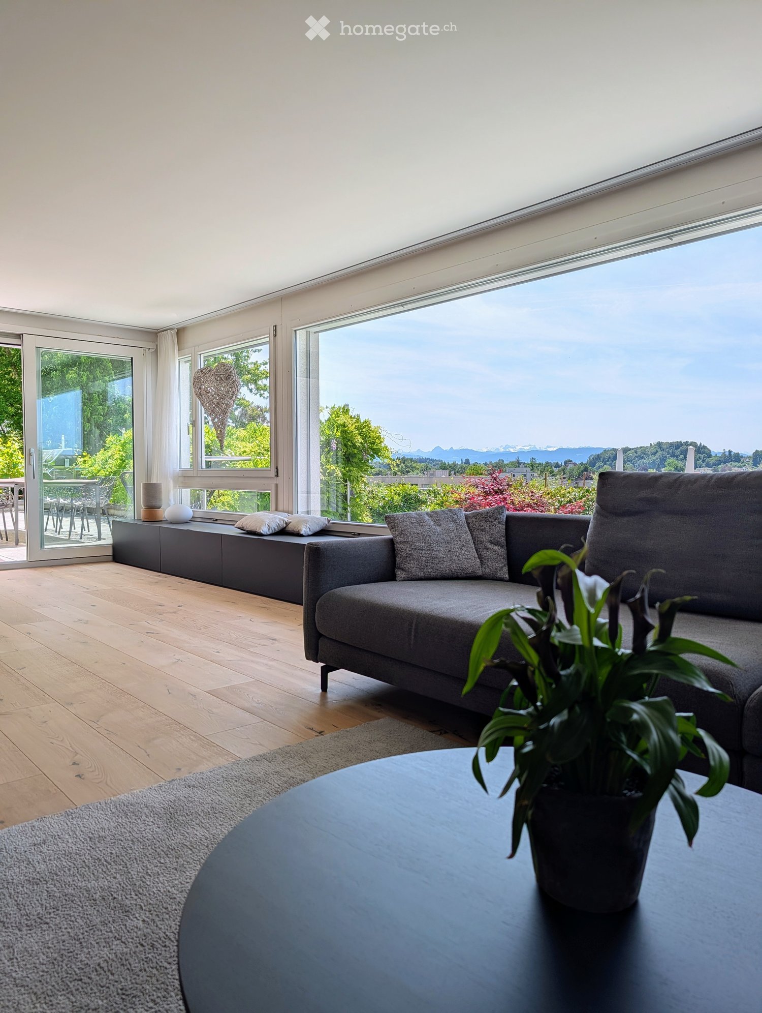Modern living room with large windows offering a scenic view, featuring a grey sofa, coffee table, potted plant, and side table.