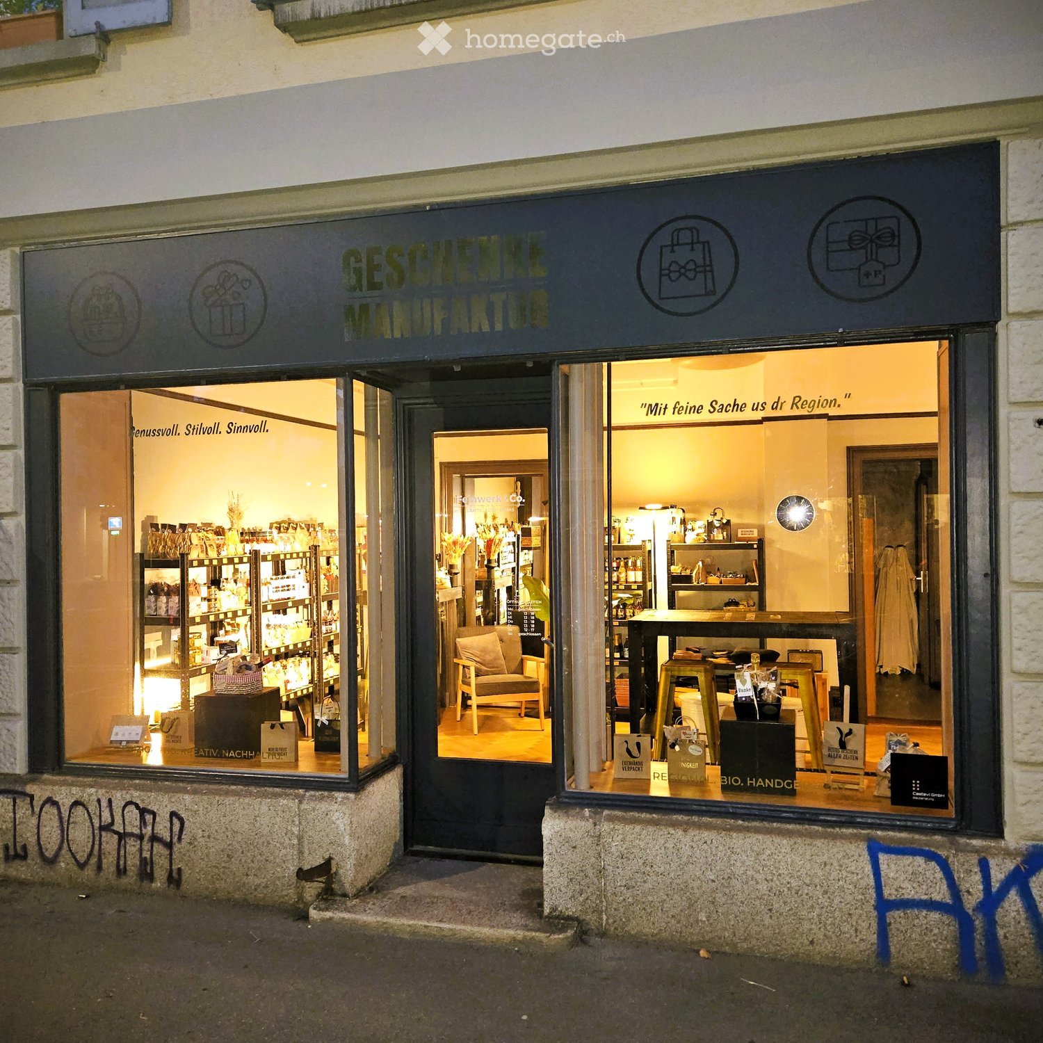 Storefront with large glass windows displaying interior shelves, a chair, and a table. The sign reads "GESCHENKE MANUFAKTUR" and the interior is well-lit with various items on display.