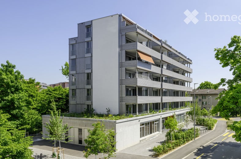 Modern multi-story residential building with clean lines, large windows, and balconies. The structure features a combination of grey and white facades, with greenery on the roof and surrounding area.