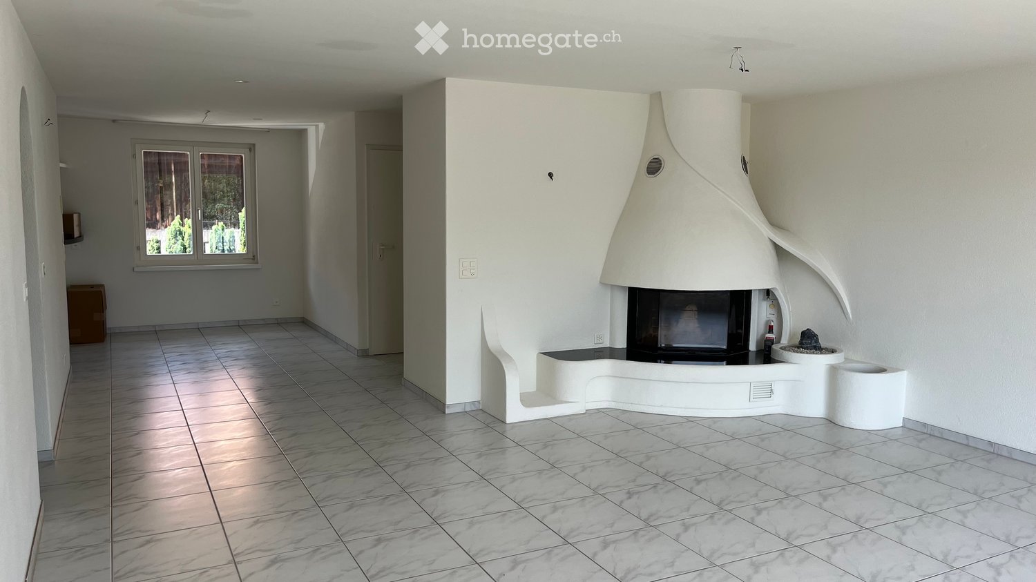 Modern living room with a white, sculptural fireplace, tiled floor, large window, and neutral-colored walls.