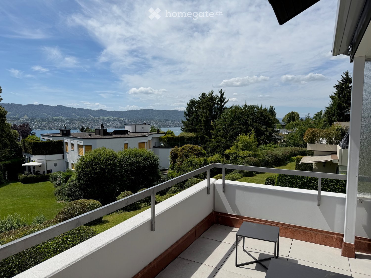 Balcony with a small table, overlooking a landscaped garden and distant lake and mountains.