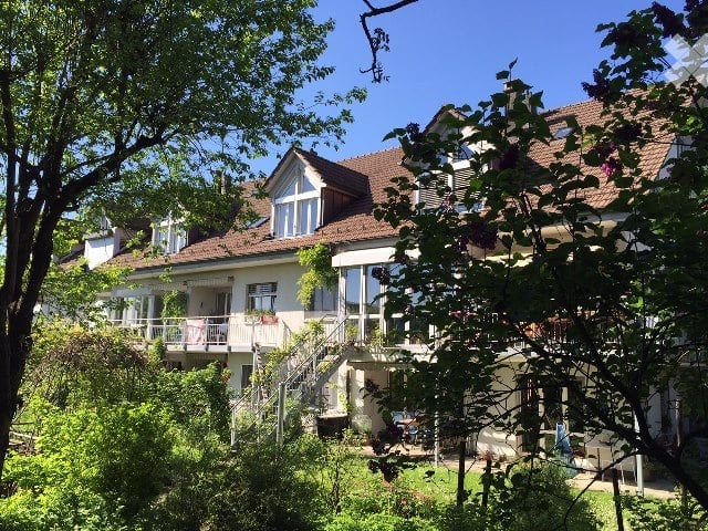A two-story house with a red-tiled roof, large windows, and a balcony. The house is surrounded by lush greenery and trees.