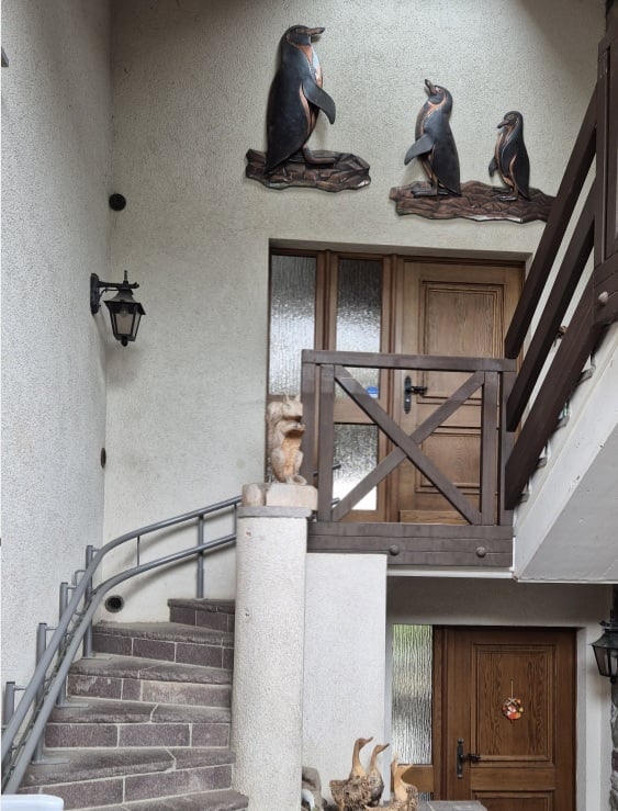 Front entrance of a house with a staircase leading to a wooden door, featuring penguin sculptures on the wall above the entrance and a small statue on the steps.
