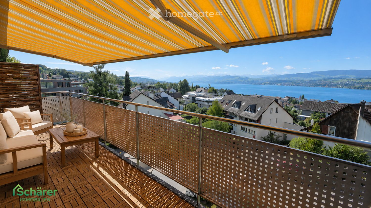 Balcony with wooden decking, beige sofa, coffee table, potted plant, and a retractable awning, overlooking a scenic lake and town view.