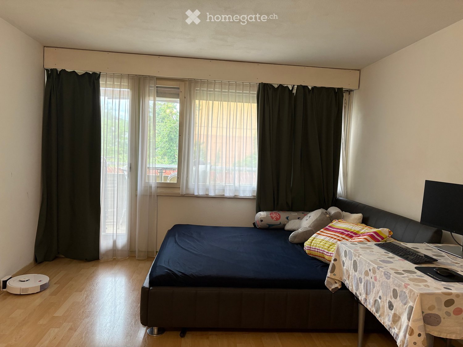 A furnished bedroom with a bed, sofa, table, laptop, and various pillows. The room features wooden flooring and large windows with curtains.