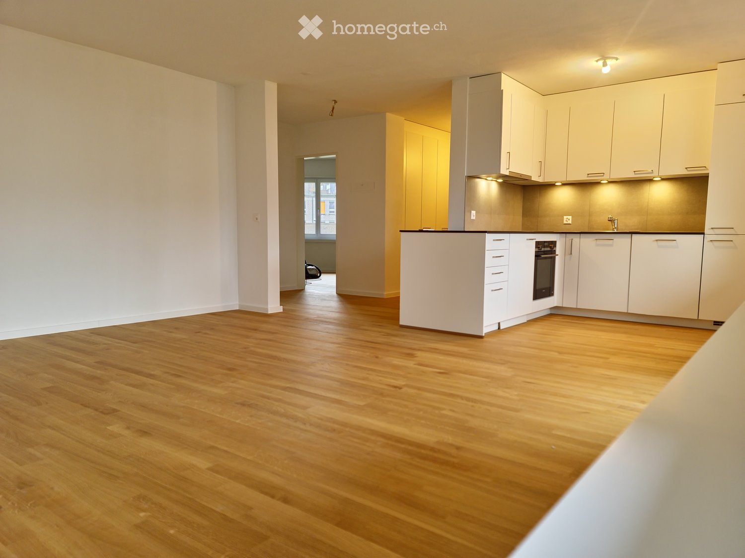 Empty room with light wooden flooring, white walls, and a small kitchen area with white cabinets and appliances.