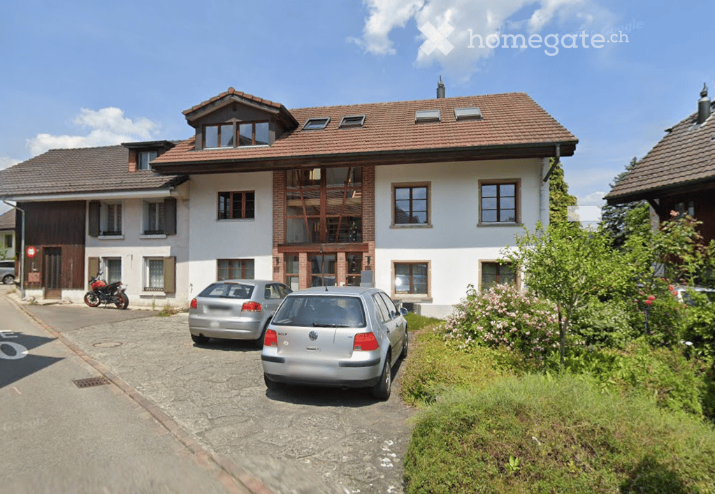 Two-story house with a red-tiled roof, white walls, and large windows. Features a garage and parking area with several cars.