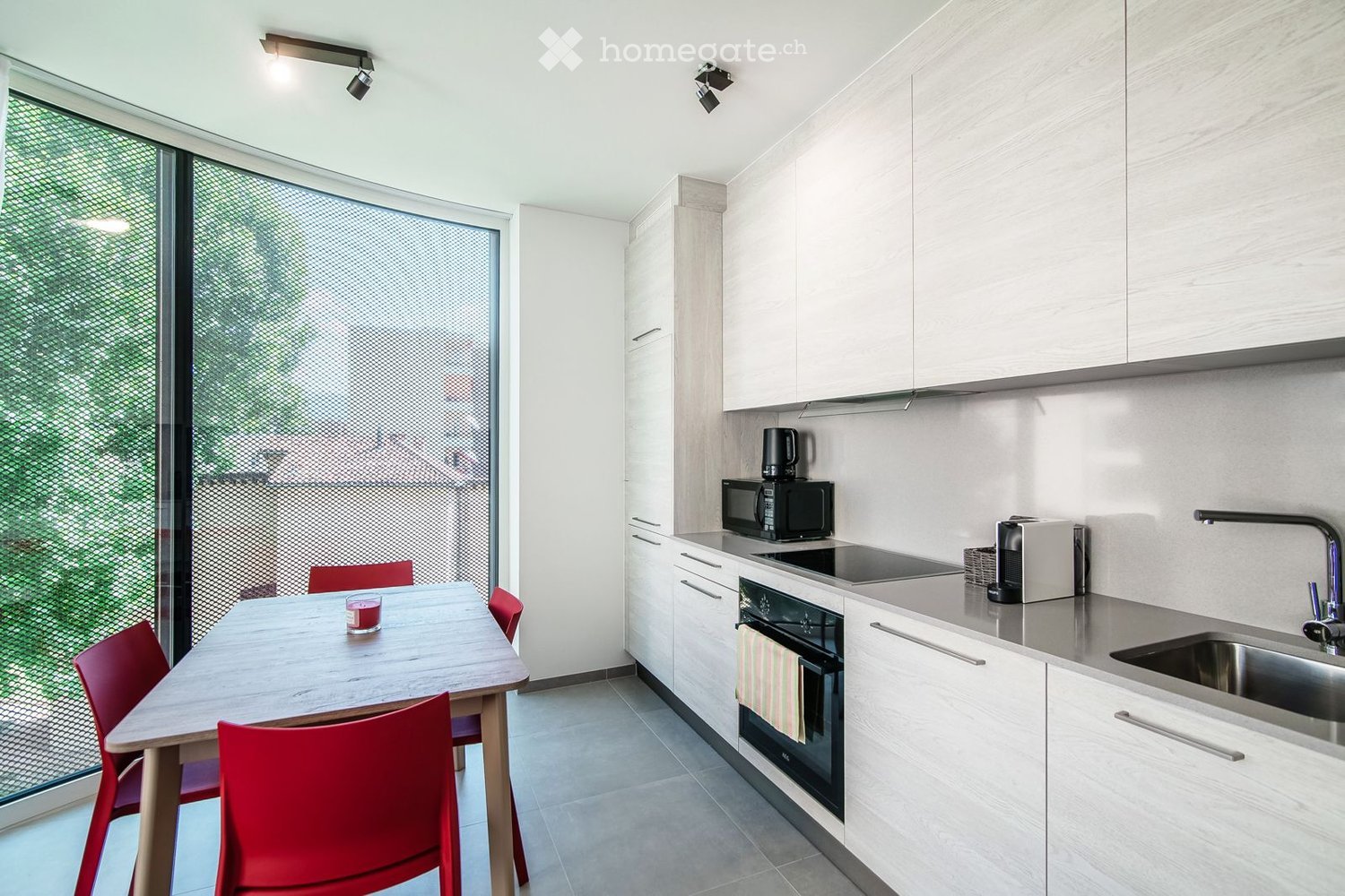 Modern kitchen with white cabinets, stainless steel appliances, a wooden dining table with red chairs, and a large window with a view of greenery.