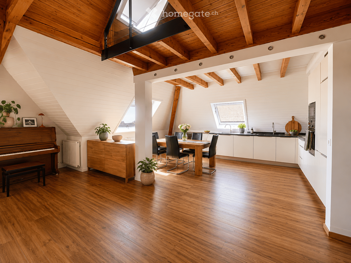 Open-plan living and dining area with wooden floors, exposed wooden beams, and white walls. Features a piano, dining table with chairs, kitchen area with cabinets and appliances, and decorative plants.