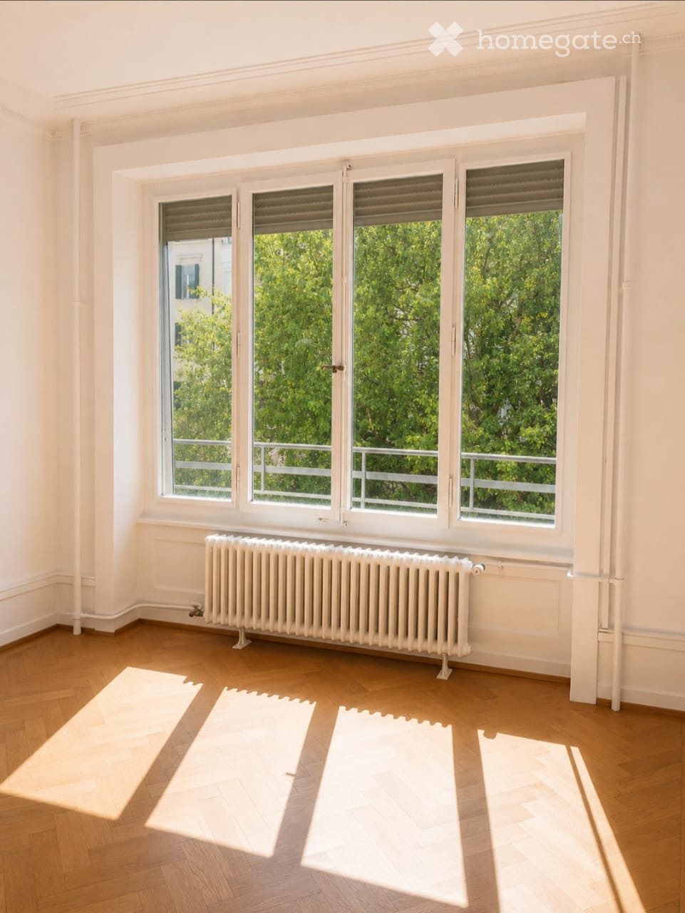 Empty room with wooden flooring, white walls, large windows with blinds, radiator, and natural light from the windows.