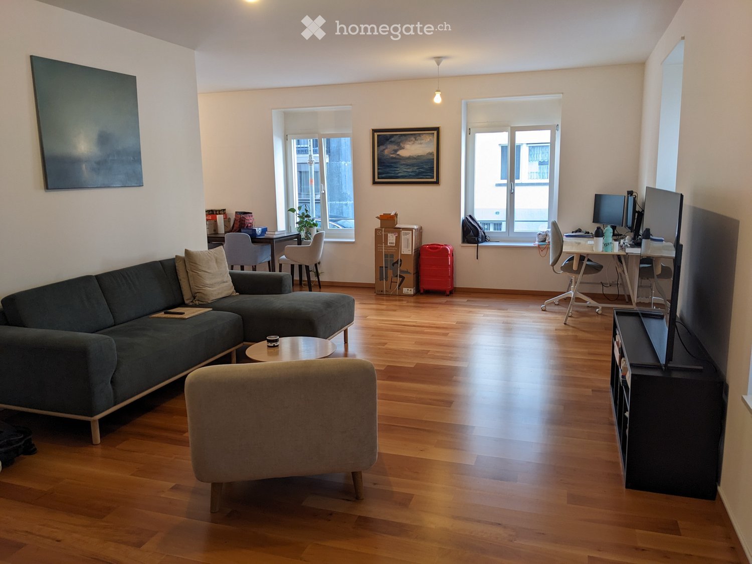 A modern living room with a dark green sectional sofa, a round coffee table, a grey armchair, a TV stand with a screen, a desk with a laptop, and a small table with a plant. The room has wooden flooring and large windows with blinds.