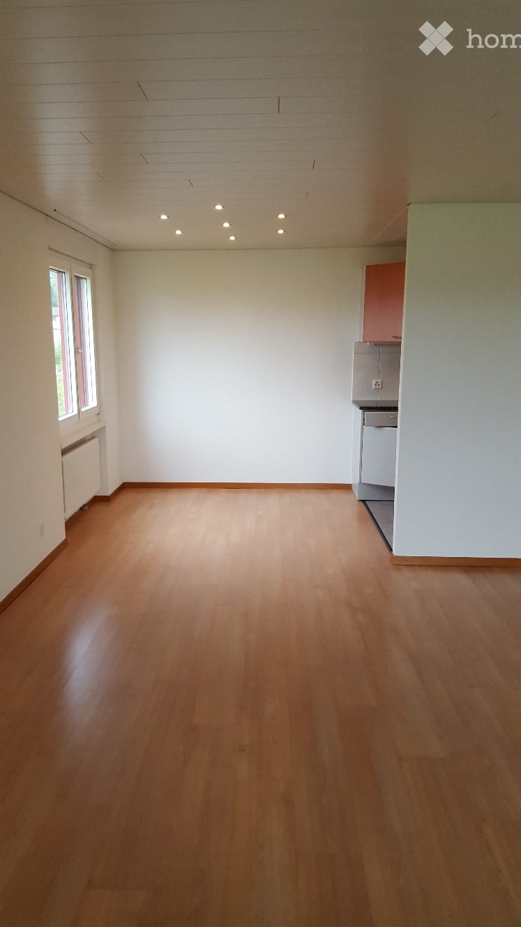 Empty room with wooden flooring, white walls, and a small kitchen area in the corner.